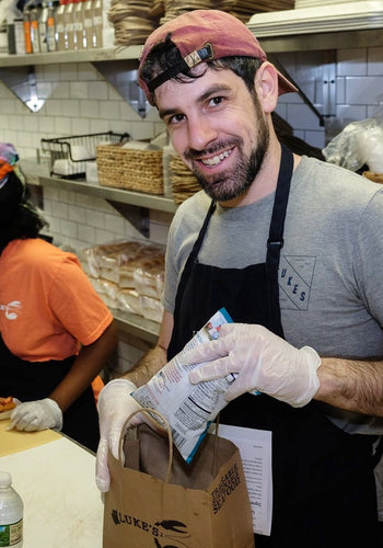 A smiling man in a professional kitchen packing a Luke's Lobster takeout bag