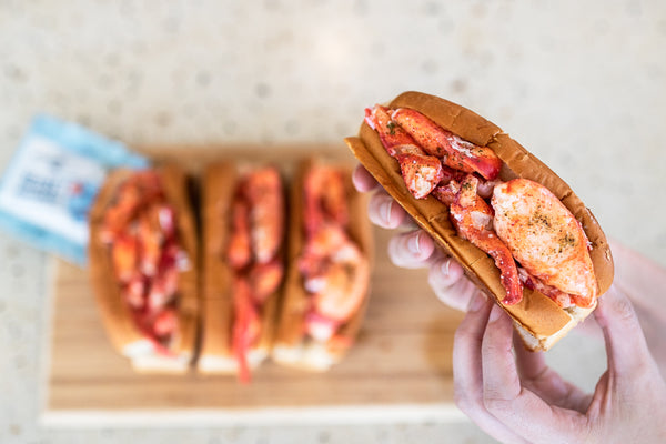 Holding up a lobster roll over a cutting board with three other lobster rolls and a package of Luke's secret seasoning