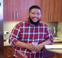 Paul Santone smiling while leaning against a kitchen table wearing a red plaid shirt