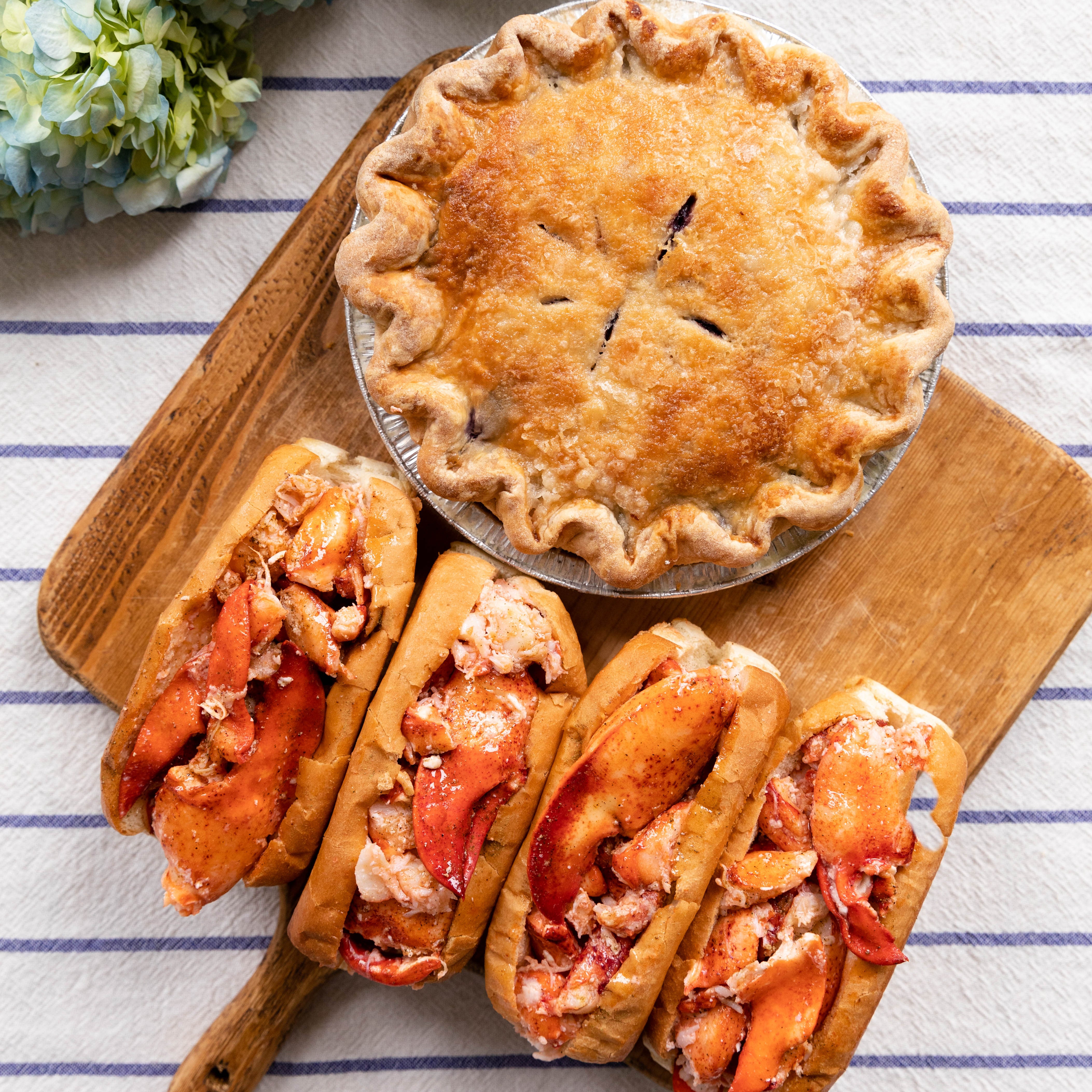 Lobster rolls and a pie on a wooden board with hydrangeas in the background