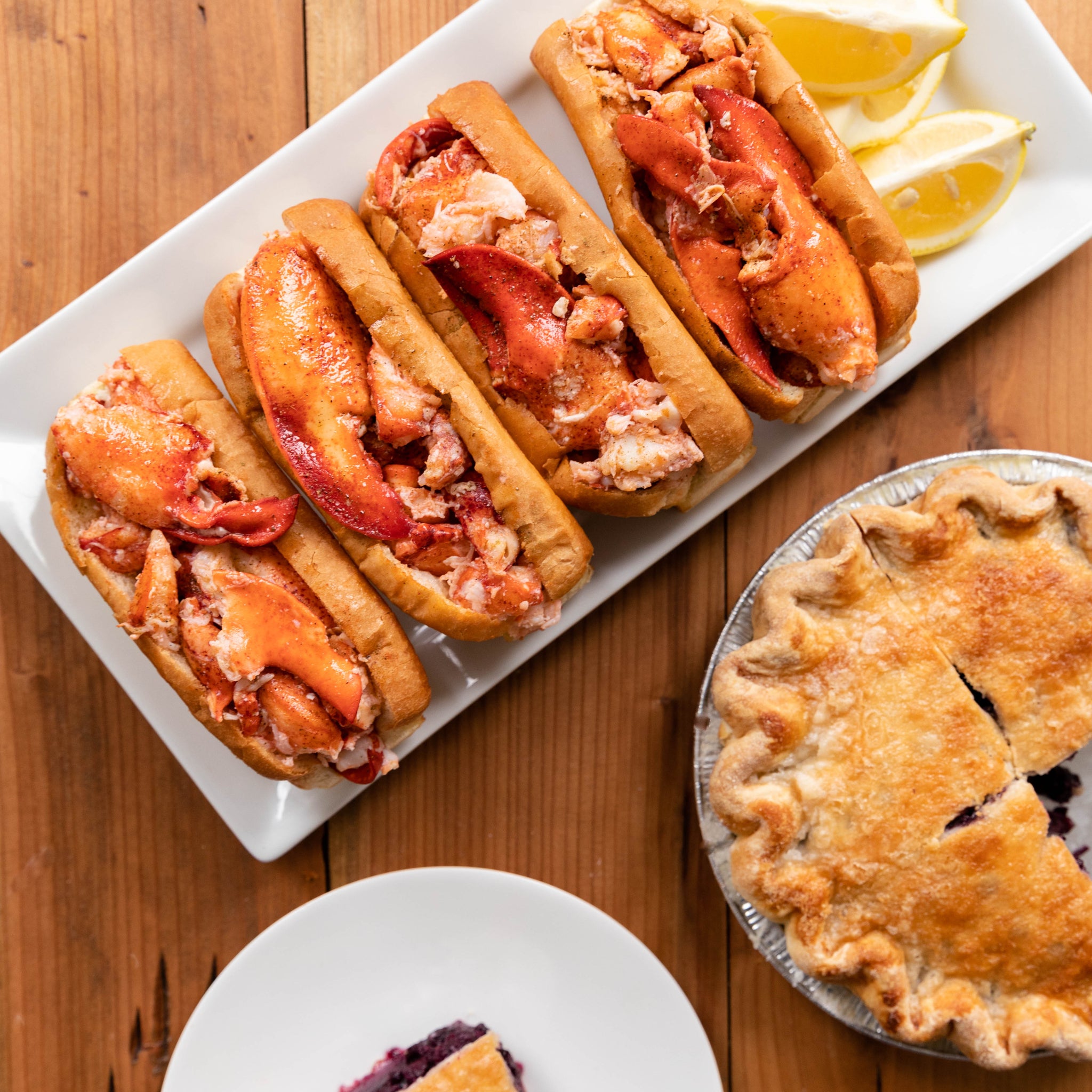 Load image into Gallery viewer, Lobster rolls on a white plate with lemon wedges, a slice of pie with ice cream, and a whole pie on a wooden table.
