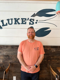 Sean; smiling in front of Luke's logo wearing a salmon shirt