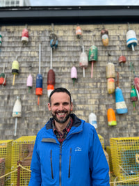 Jim wearing a blue rain jacket posing and smiling in front of a wall of buoys and a row of lobster traps.