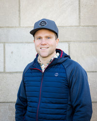 Adam smiling while wearing a blue hat, a navy jacket, and a plaid button up