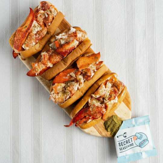 Three lobster rolls on a wooden board with a packet of 'Secret Seasoning' on a light background