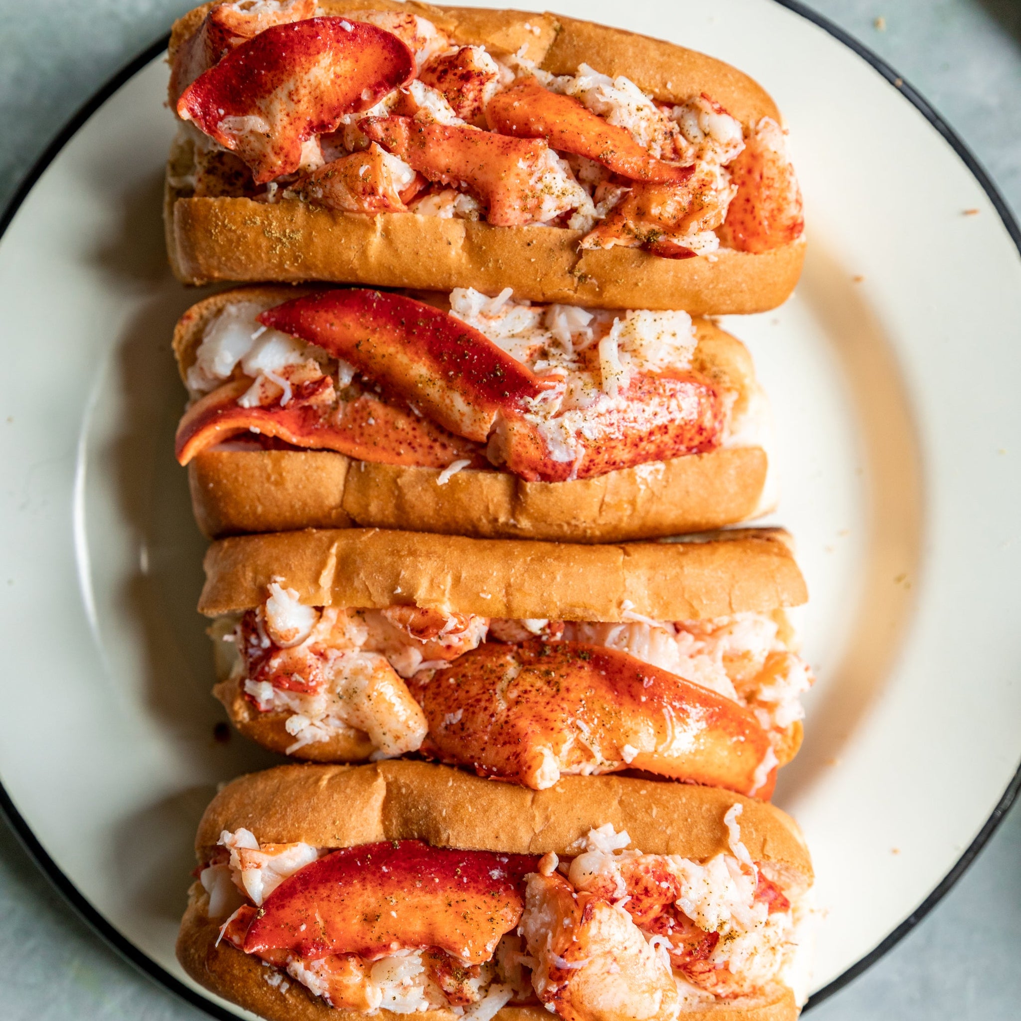 Load image into Gallery viewer, Three lobster rolls stacked on a white plate with a black rim against a light blue background.
