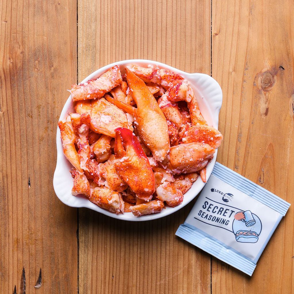 Load image into Gallery viewer, A white bowl filled with fresh lobster meat, accompanied by a packet of seasoning blend, placed on a wooden surface.
