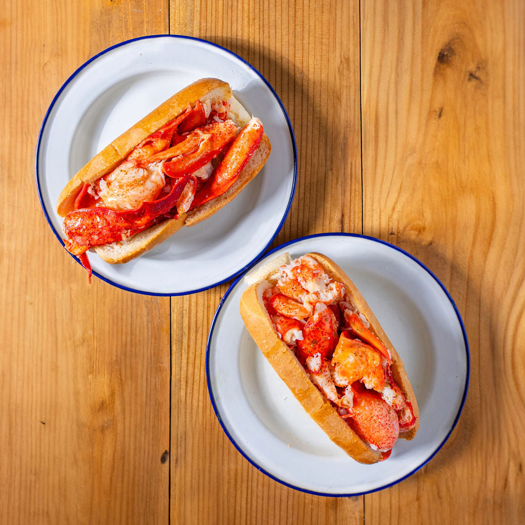 Load image into Gallery viewer, Two lobster rolls on white plates with blue rims on a wooden surface
