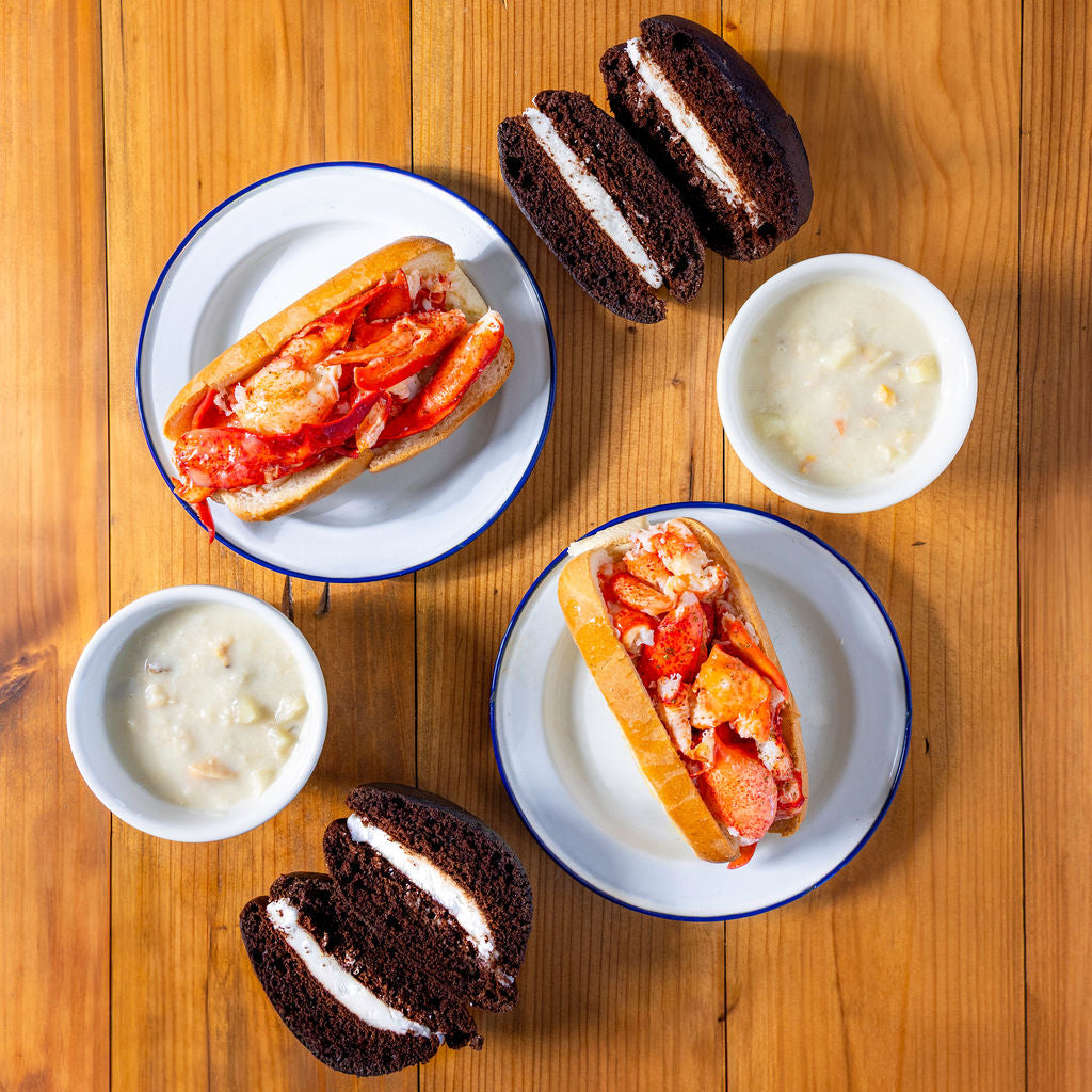 Load image into Gallery viewer, Two lobster rolls on white plates with blue rims, accompanied by two small bowls of soup on a wooden surface.
