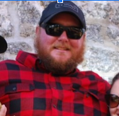 Bryan wearing a flannel, sunglasses, and baseball hat smiling for a photo